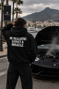Color Black — OVRSAID BE REALISTIC: EXPECT A MIRACLE. oversized hoodie on male model, back view showing the white back print
